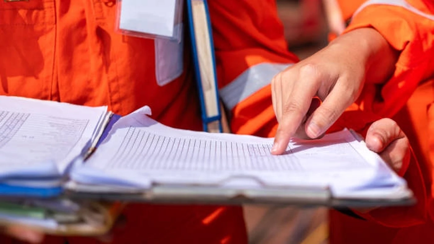 A worker in orange coveralls reviews entries in an open field log while a colleague points at a line on the page.